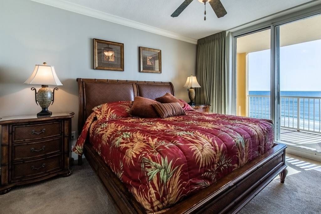 A large bed with a floral bedspread, two bedside lamps, a wooden nightstand, and a view of the ocean through a balcony.
