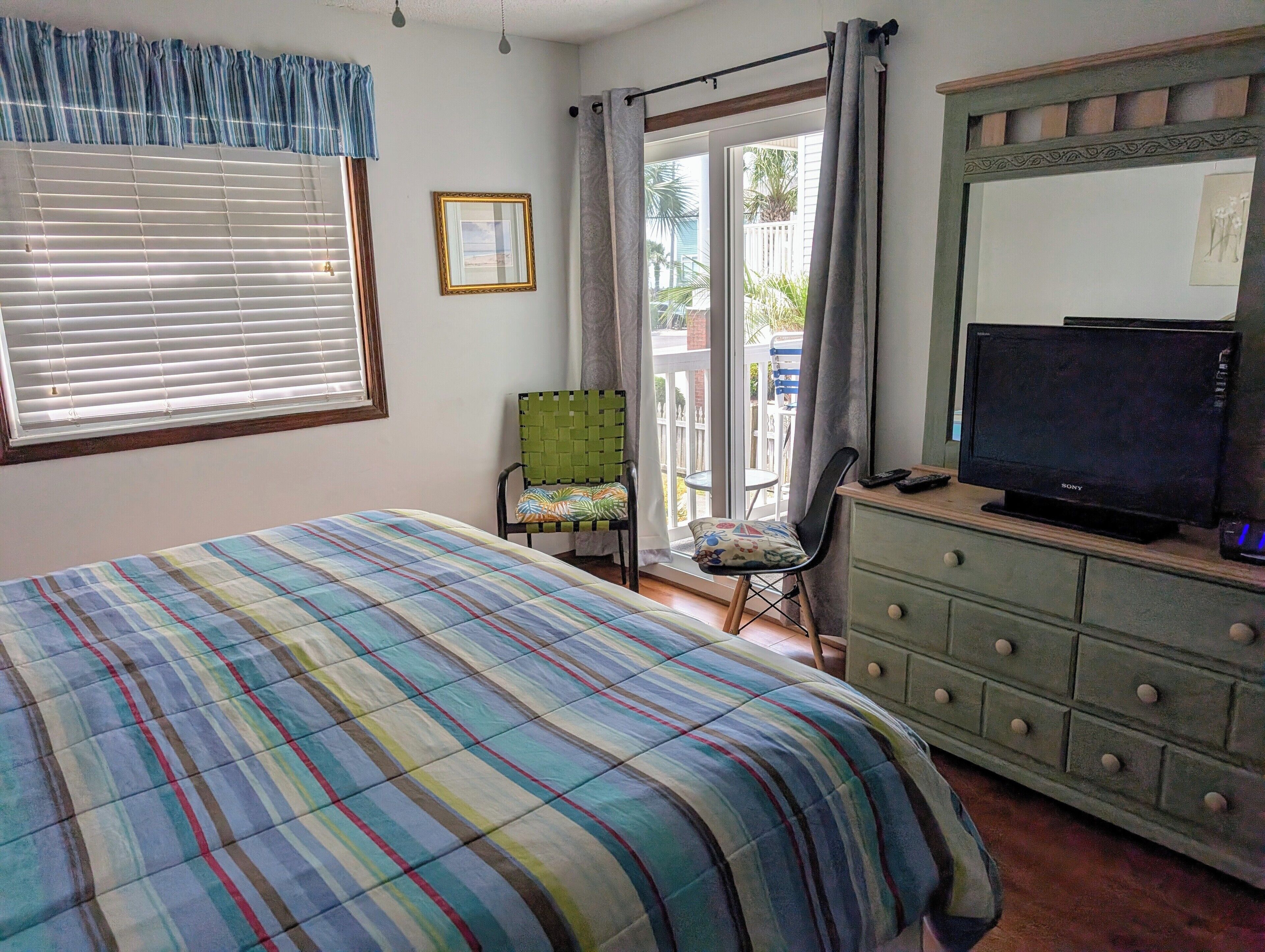 A bedroom with a bed, a chair, a dresser, a television, and a window with blinds.