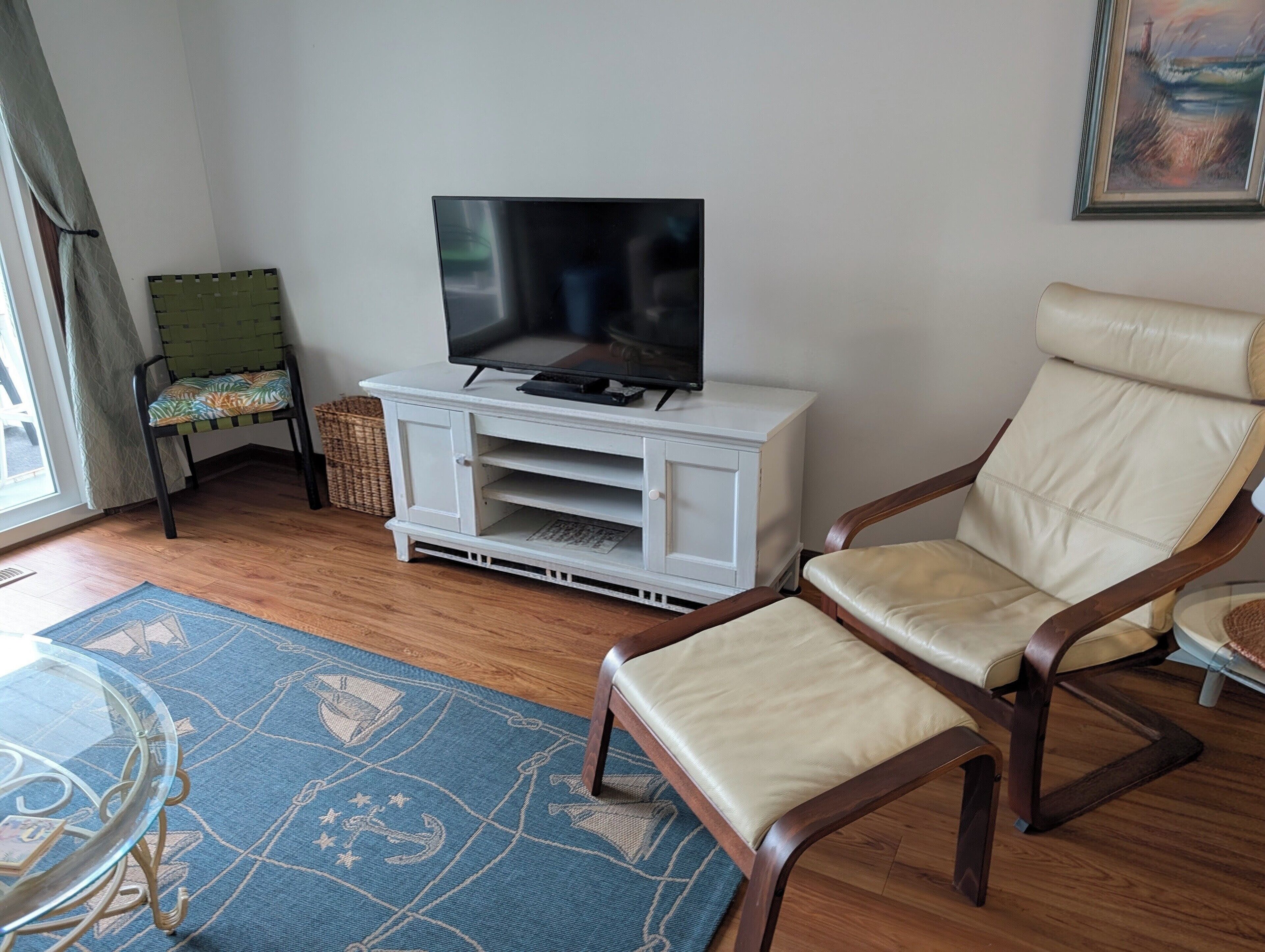 A living room with a television on a white cabinet, a beige leather chair, a patterned rug, and a wicker basket.