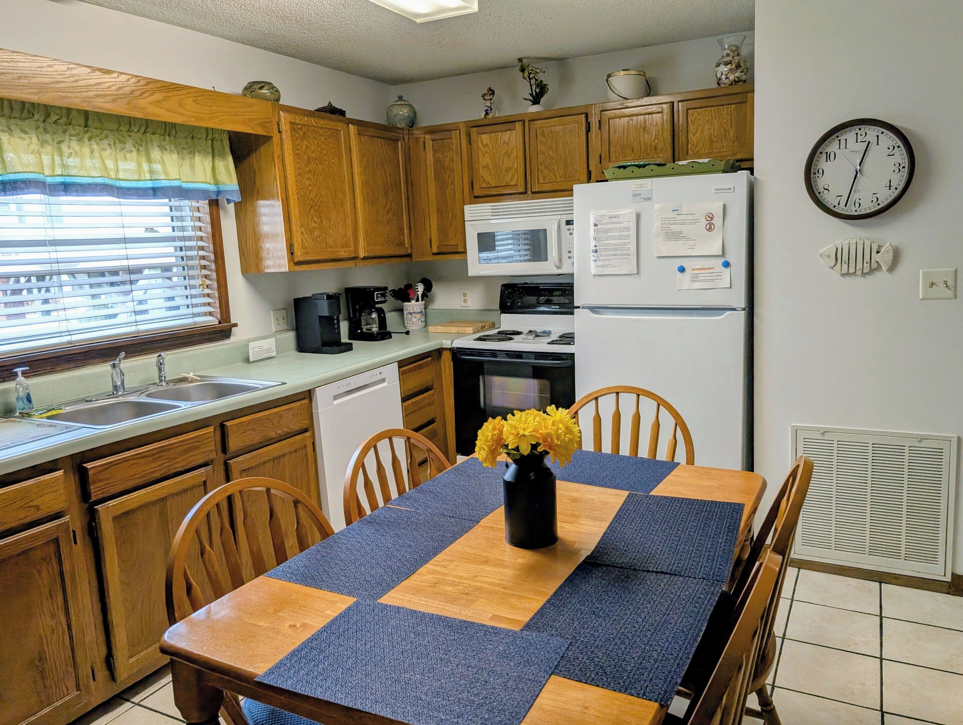 Une cuisine avec des meubles de rangement en bois, un four à micro-ondes, une cuisinière, un réfrigérateur et un évier. Il y a une horloge au mur et un vase avec des fleurs sur la table.