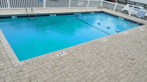 Una piscina rettangolare con acqua cristallina blu, circondata da una recinzione bianca. Sono presenti indicatori di profondità che segnalano 3, 4 e 6 piedi. La piscina è adiacente a una zona pavimentata con motivo a mattoni.