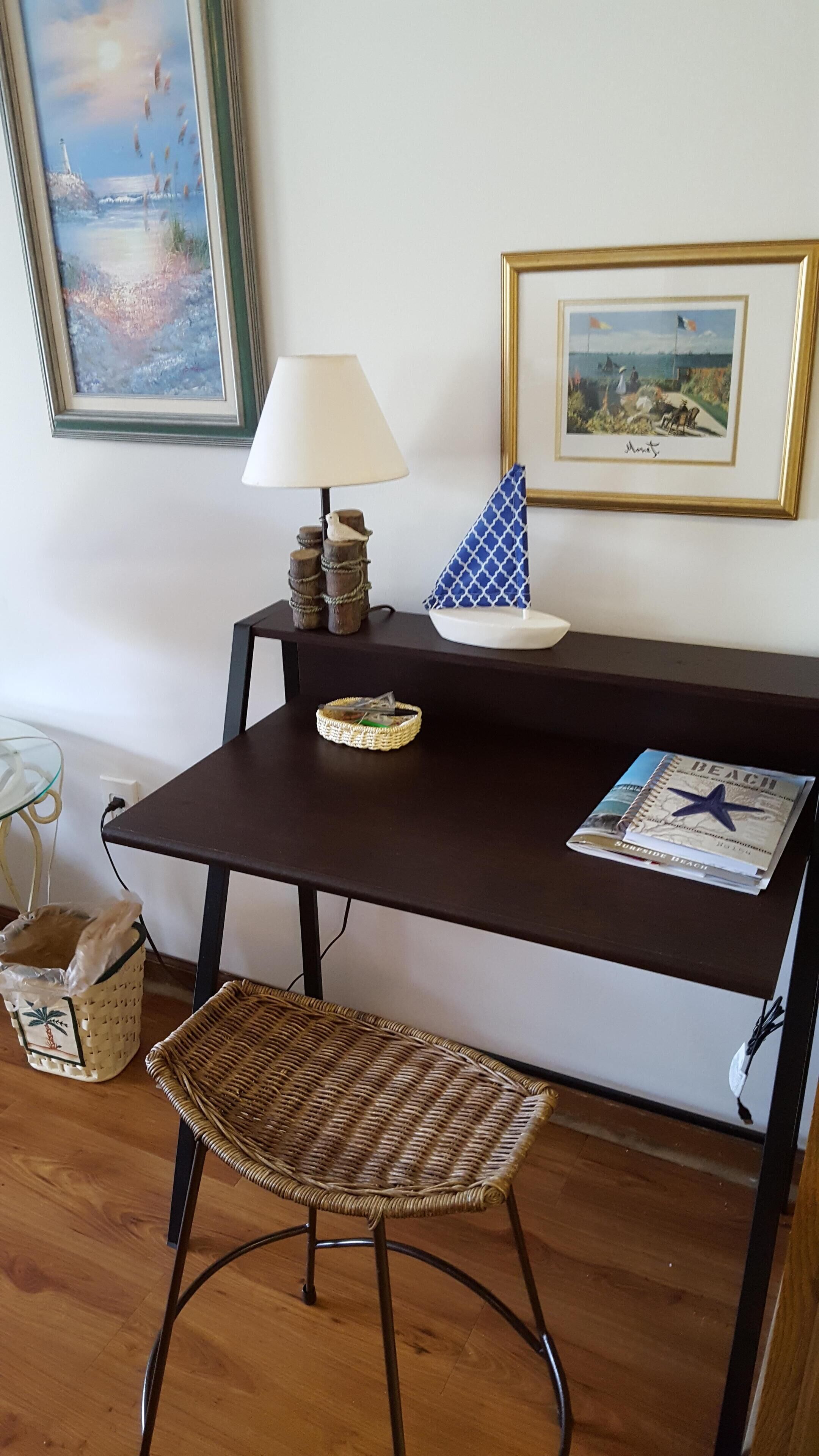 A dark brown desk with a lamp, a wicker chair, a framed painting, and a beach-themed magazine.