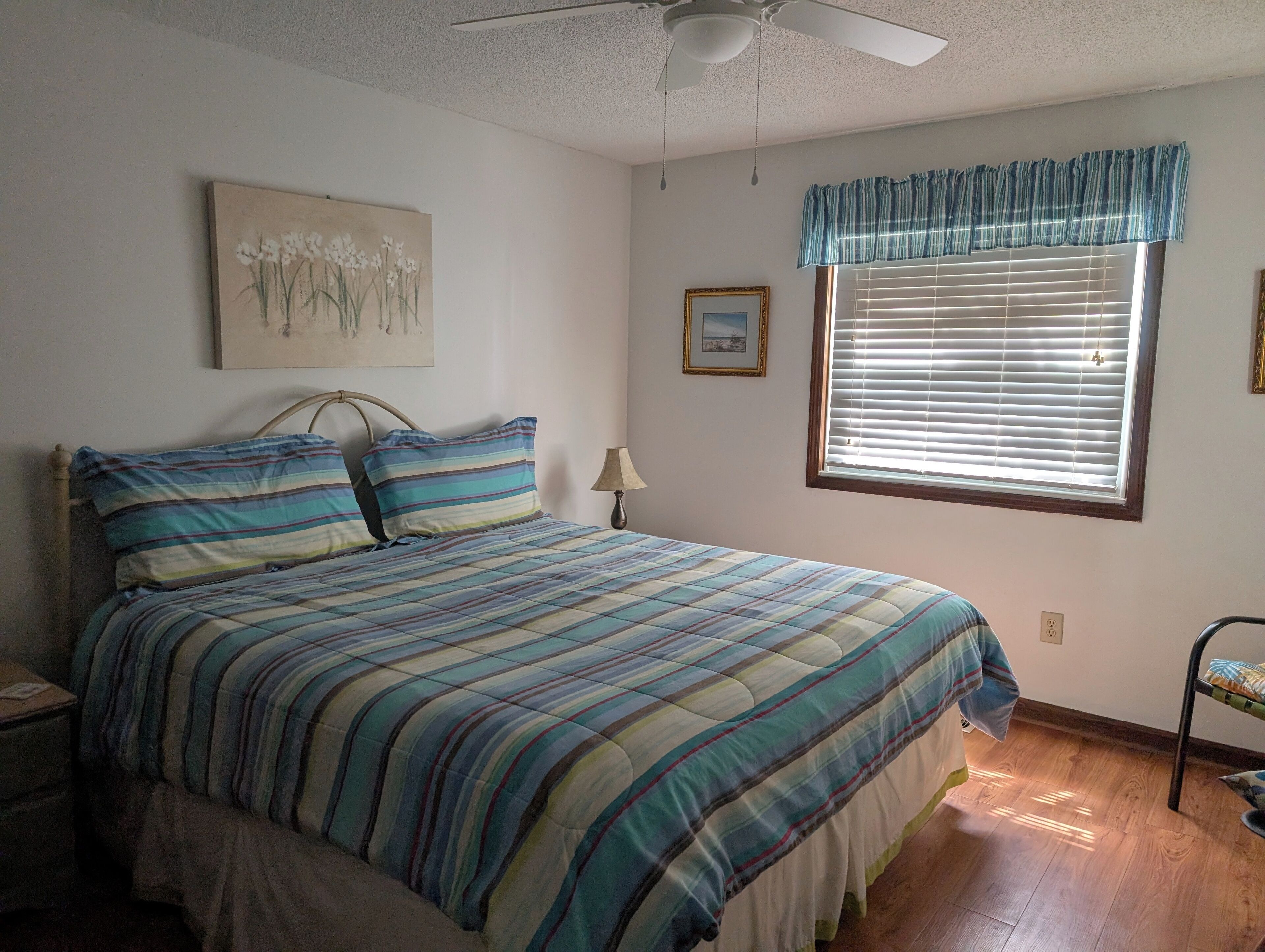 Un dormitorio con una cama, un ventilador de techo, una ventana con persianas, una lámpara y un cuadro en la pared.