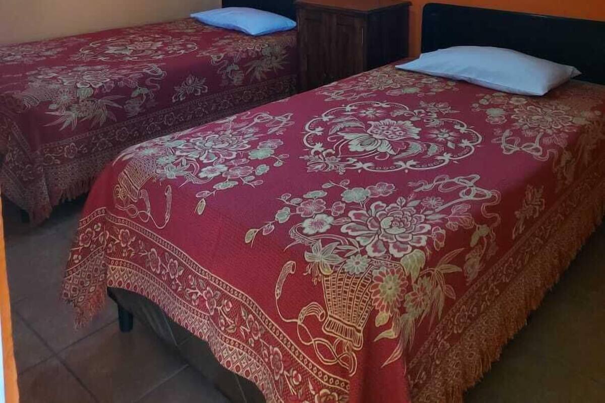 A room with two beds, each with a floral-patterned bedspread and a pillow. There are two windows on the orange wall, and the ceiling is wooden.