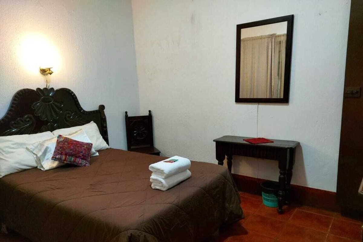 A neatly made bed with a brown bedspread and white pillows, a small table with a red napkin, a wall-mounted mirror, and a trash bin.