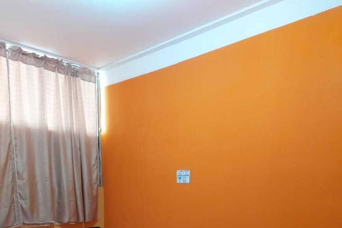 A room with an orange wall, a bed with a red bedspread, a window with beige curtains, and a small sign on the wall.