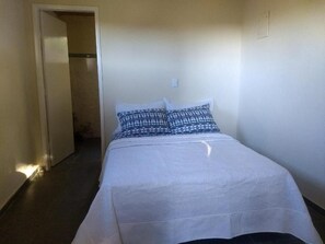 A single bed with a white bedspread and patterned pillows in a room with plain walls and a door leading to a bathroom. - Sítio Colibri - Serra da Mantiqueira (Brazópolis)