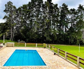 A rectangular swimming pool with a stone tiled deck, surrounded by a wooden fence and a grassy area with trees in the background. - Sítio Colibri - Serra da Mantiqueira (Brazópolis)