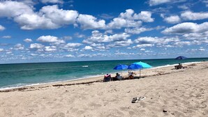 Ein Strand mit blauem Himmel und ruhigem Meer. Menschen entspannen sich unter blauen Sonnenschirmen.