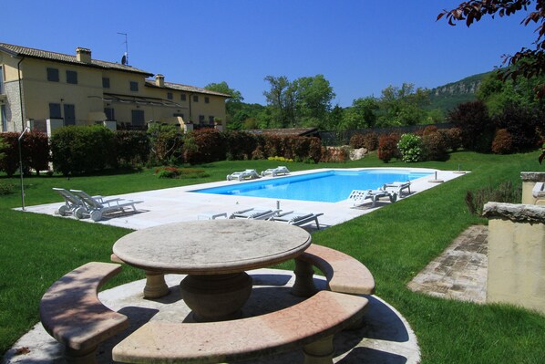 Uma área de piscina com mesa e cadeiras, cercada por um gramado bem cuidado e um edifício ao fundo.