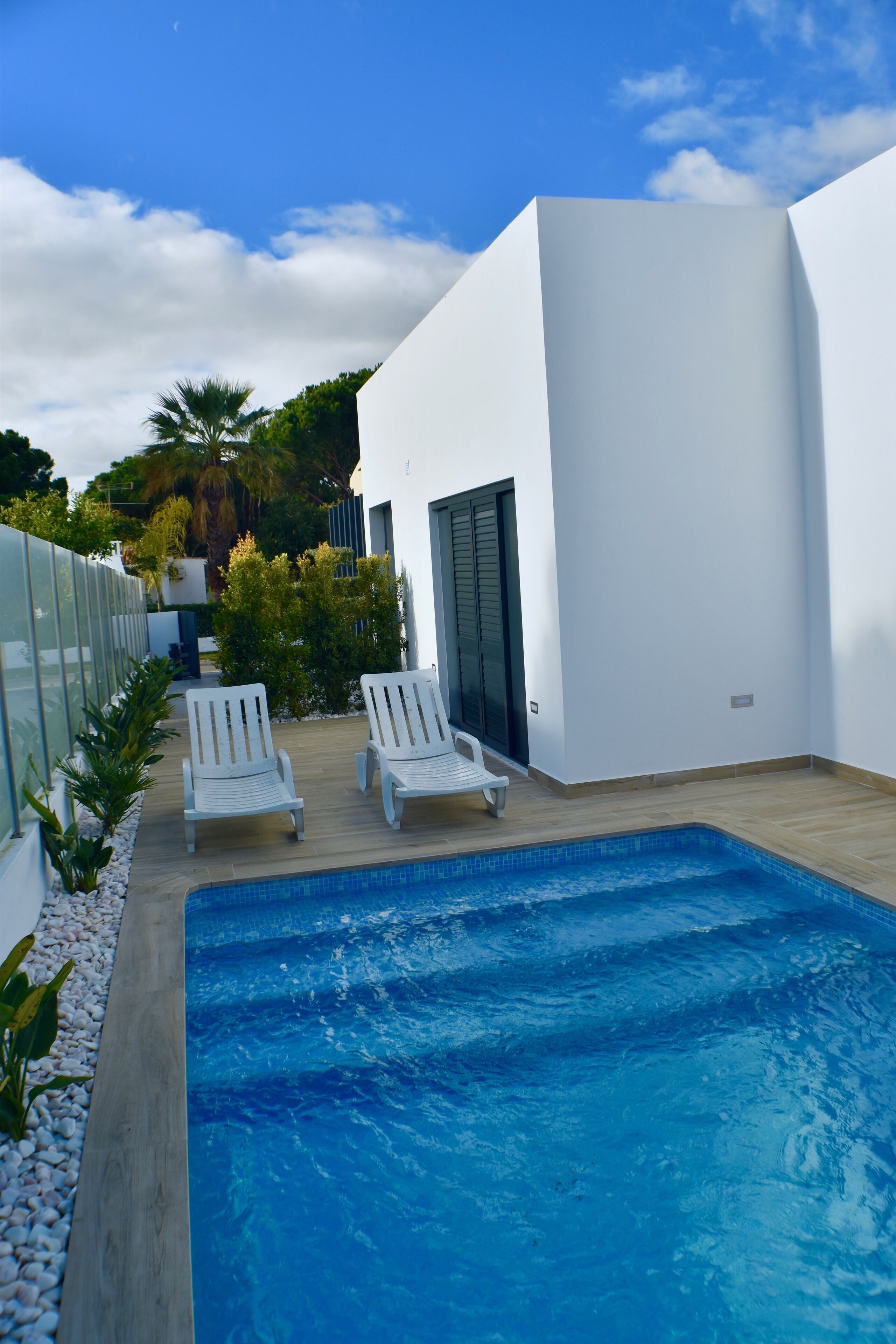 A modern outdoor pool area with two white lounge chairs, a wooden deck, and a white building with a door.