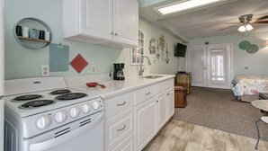 Une cuisine compacte avec des meubles blancs, une cuisinière et une machine à café. On y trouve une petite étagère ronde avec des livres et un objet rouge posé sur le comptoir.