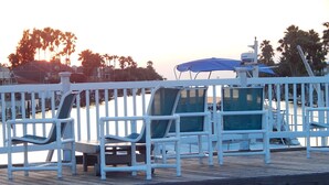Une terrasse en bois avec des balustrades blanches, deux parasols bleus et un bateau en arrière-plan.