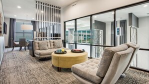 Ein modernes Hotelzimmer mit Sofa, Sesseln und Pouf, einem großen Fenster mit Blick auf die Stadt sowie einer dekorativen Wandverkleidung.