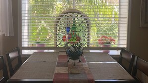 Uma área de refeições com uma mesa posta para dois, um arranjo decorativo e vista para o verde através de uma janela com persianas.