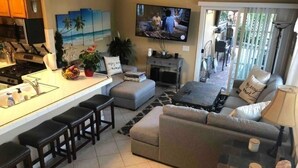 Uma cozinha moderna com ilha central, área de jantar com sofá e cadeiras, e sala de estar com uma TV grande.