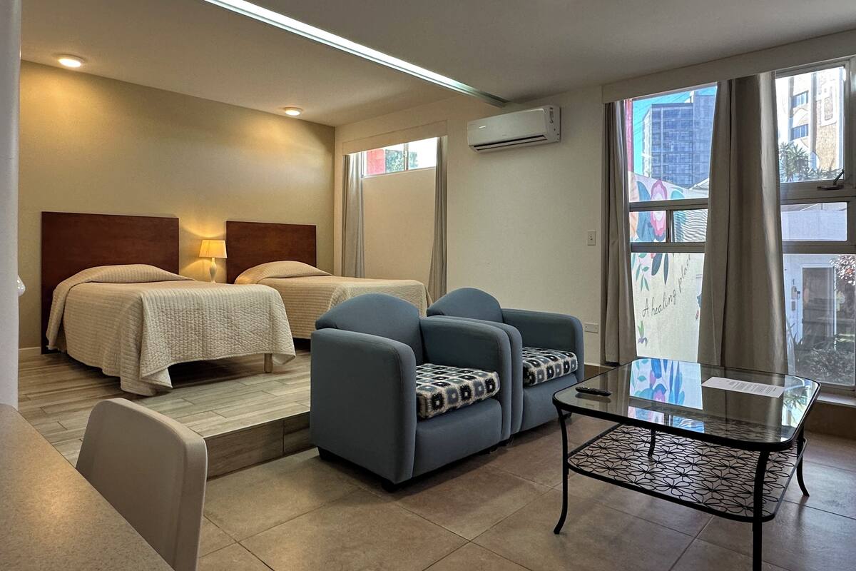 A hotel room with two beds, a sofa, a glass coffee table, and a window with a view of buildings.