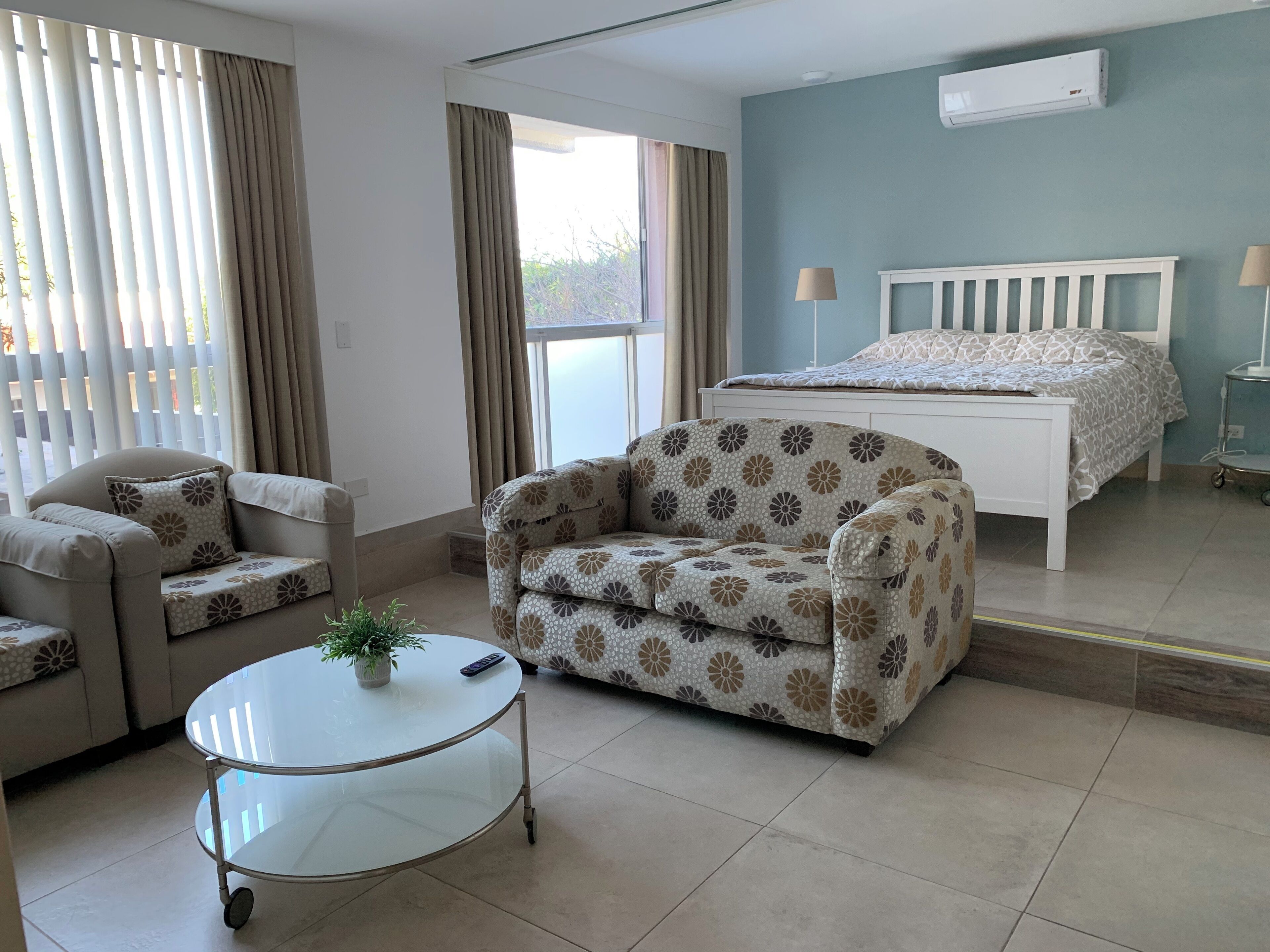 A modern room with a sofa, a round glass table with a plant, a bed with a patterned bedspread, and a window with curtains.