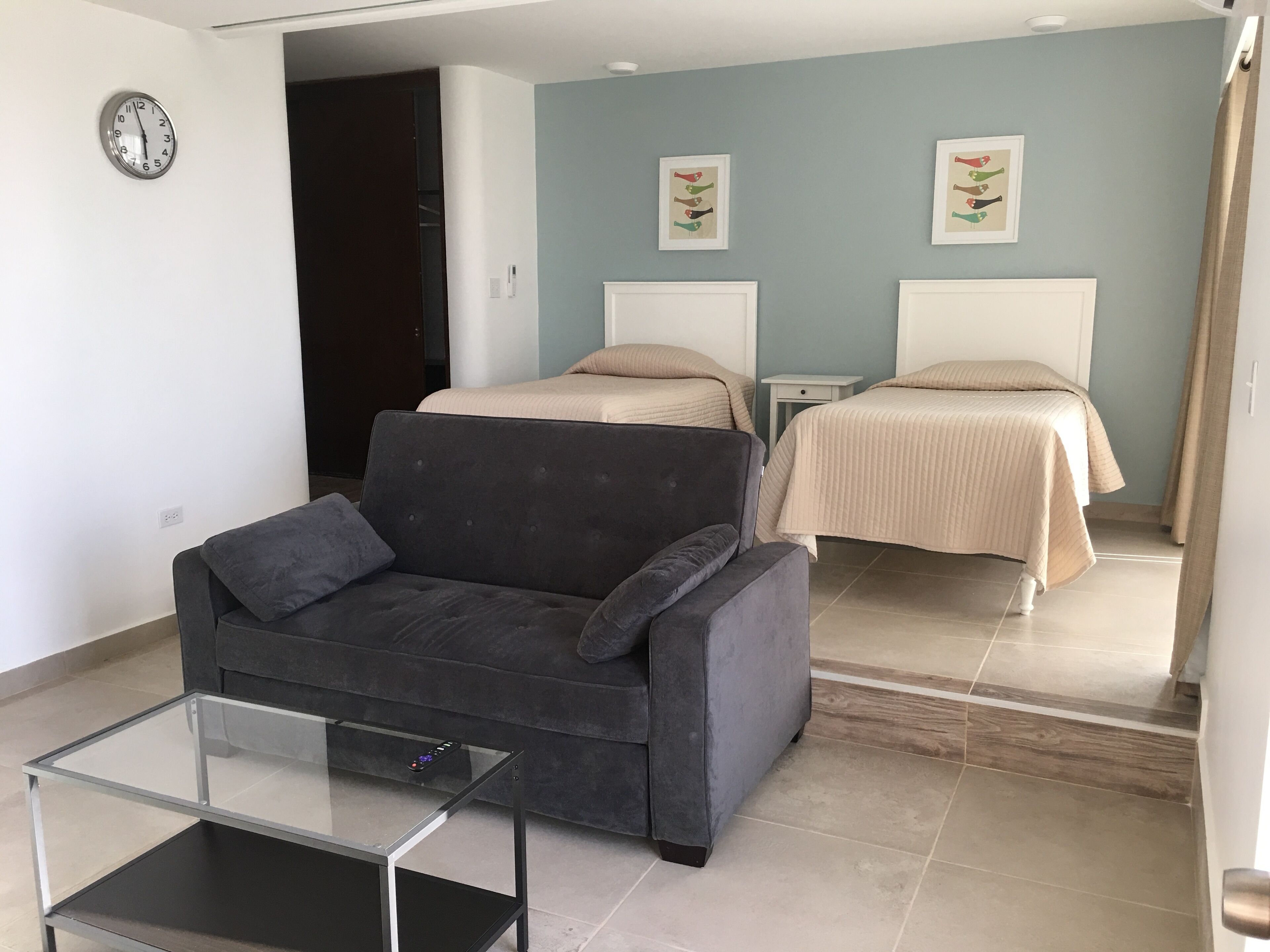 A modern room with a grey sofa, a glass-top coffee table, two beds with beige bedding, and a wall clock.