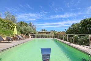 Una piscina con sillas de descanso y sombrillas, rodeada de vegetación y una pérgola.