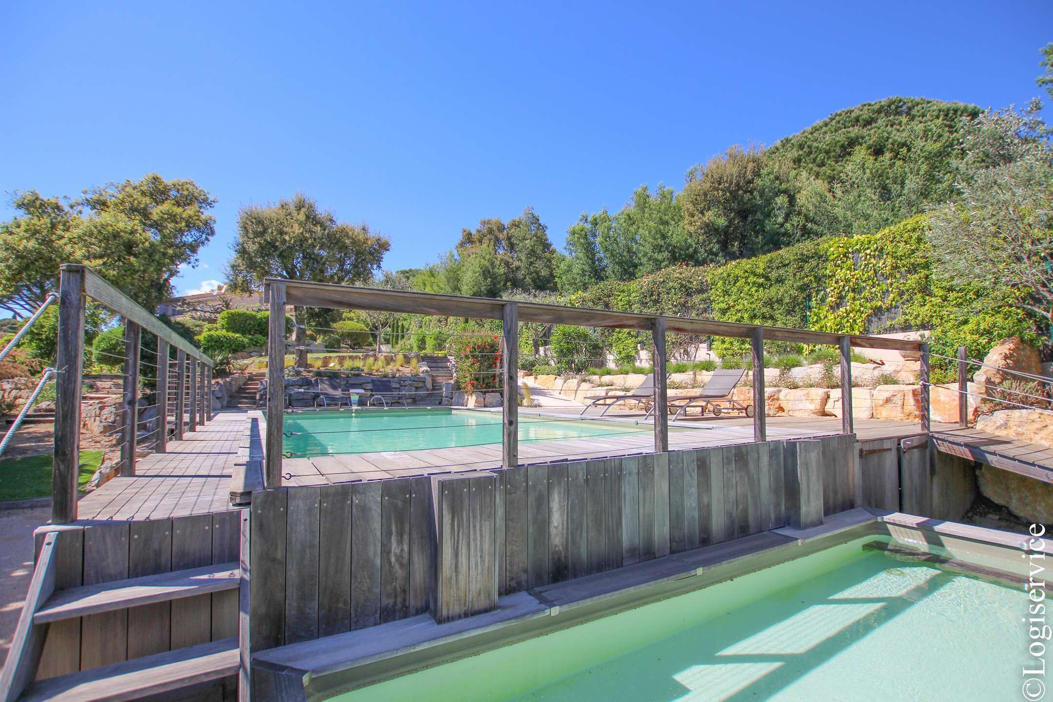 Eine Holzterrasse mit Pool und Liegestühlen, umgeben von Grünflächen und Hügeln.