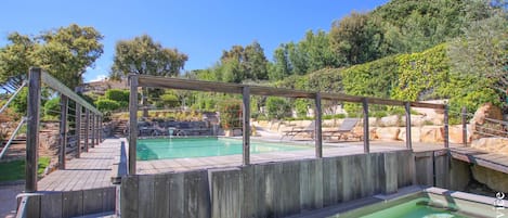 Eine Holzterrasse mit Pool und Liegestühlen, umgeben von Grünflächen und Hügeln.