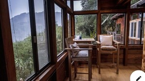 A wooden cabin with large windows, a wooden table, two chairs, and a view of a forested area. - Gorgeous Secluded Treehouse with Spectacular Views (Carmel Valley)