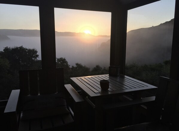 A wooden table and chairs in a covered outdoor area with a view of a sunrise over a misty landscape. - Gorgeous Secluded Treehouse with Spectacular Views (Carmel Valley)