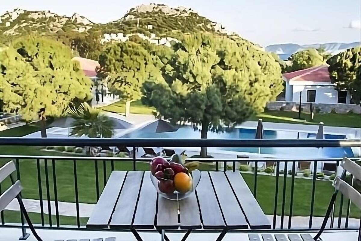 A balcony with a table and chairs, a bowl of fruit, and a view of trees and buildings.
