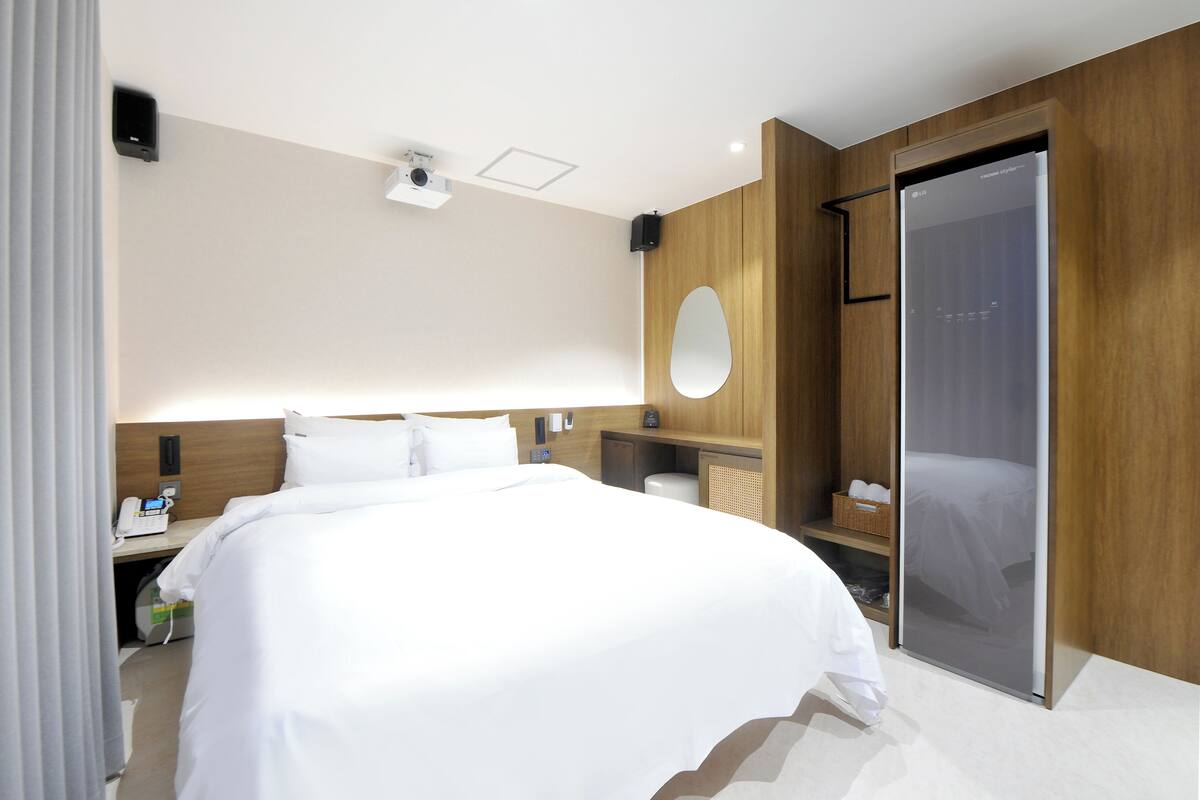 Una habitación de hotel moderna con una cama grande, un cabecero de madera, una mesita de noche con lámpara, un espejo y un armario con puertas correderas.