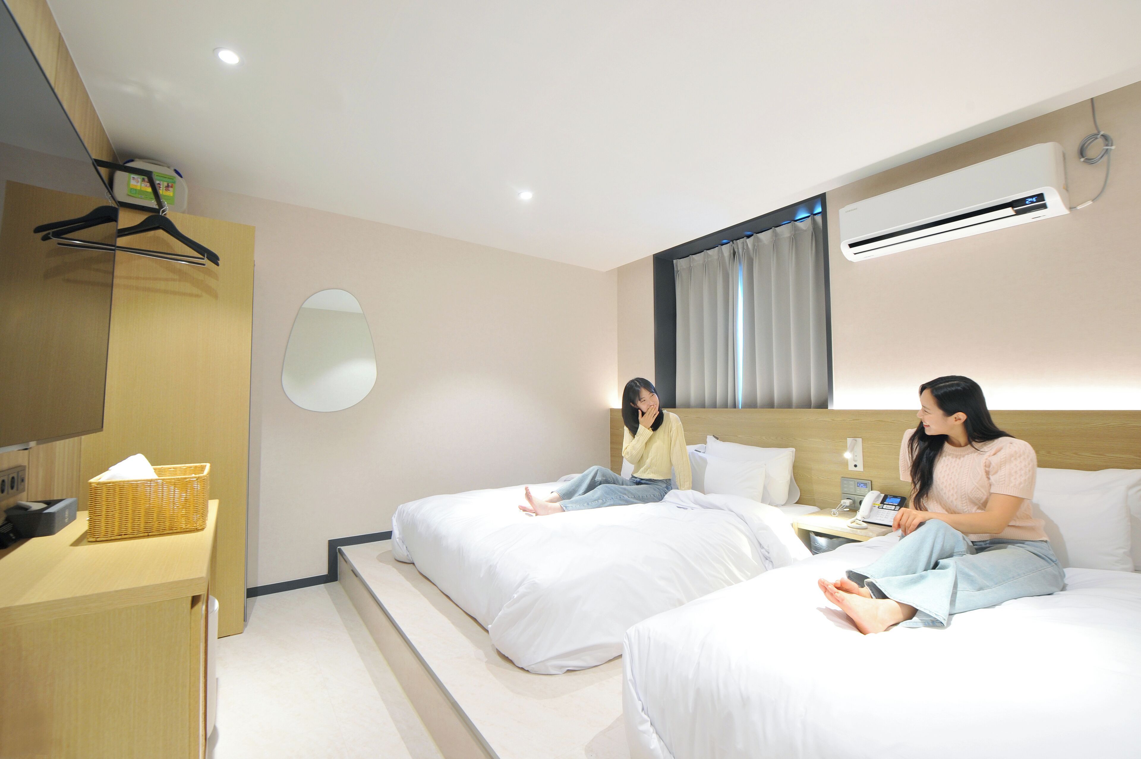 A hotel room with two beds, a desk, a basket, and an air conditioning unit.