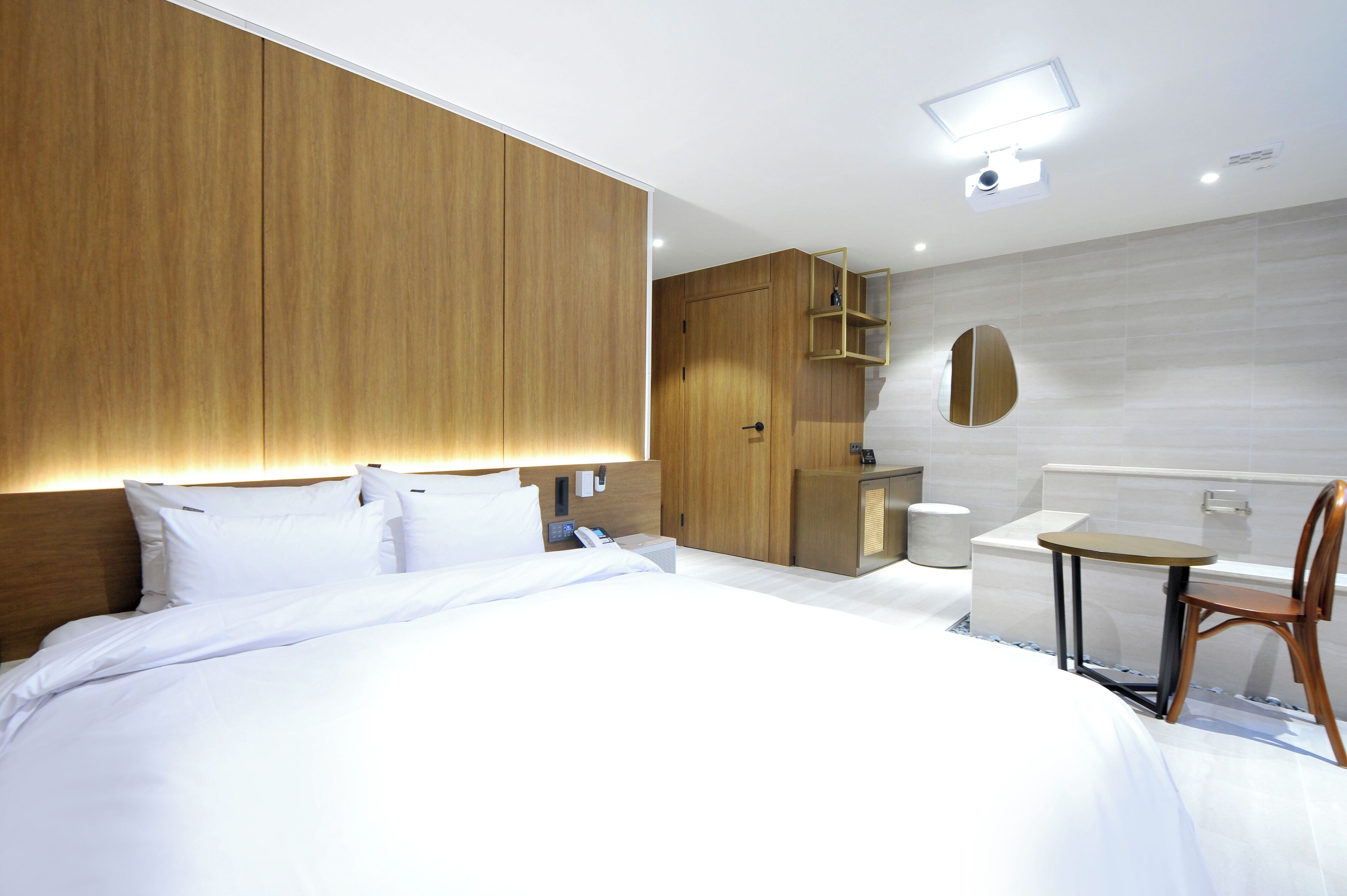 A modern hotel room with a large bed, wooden headboard, a small table with a chair, a built-in desk, and a mirror on the wall.