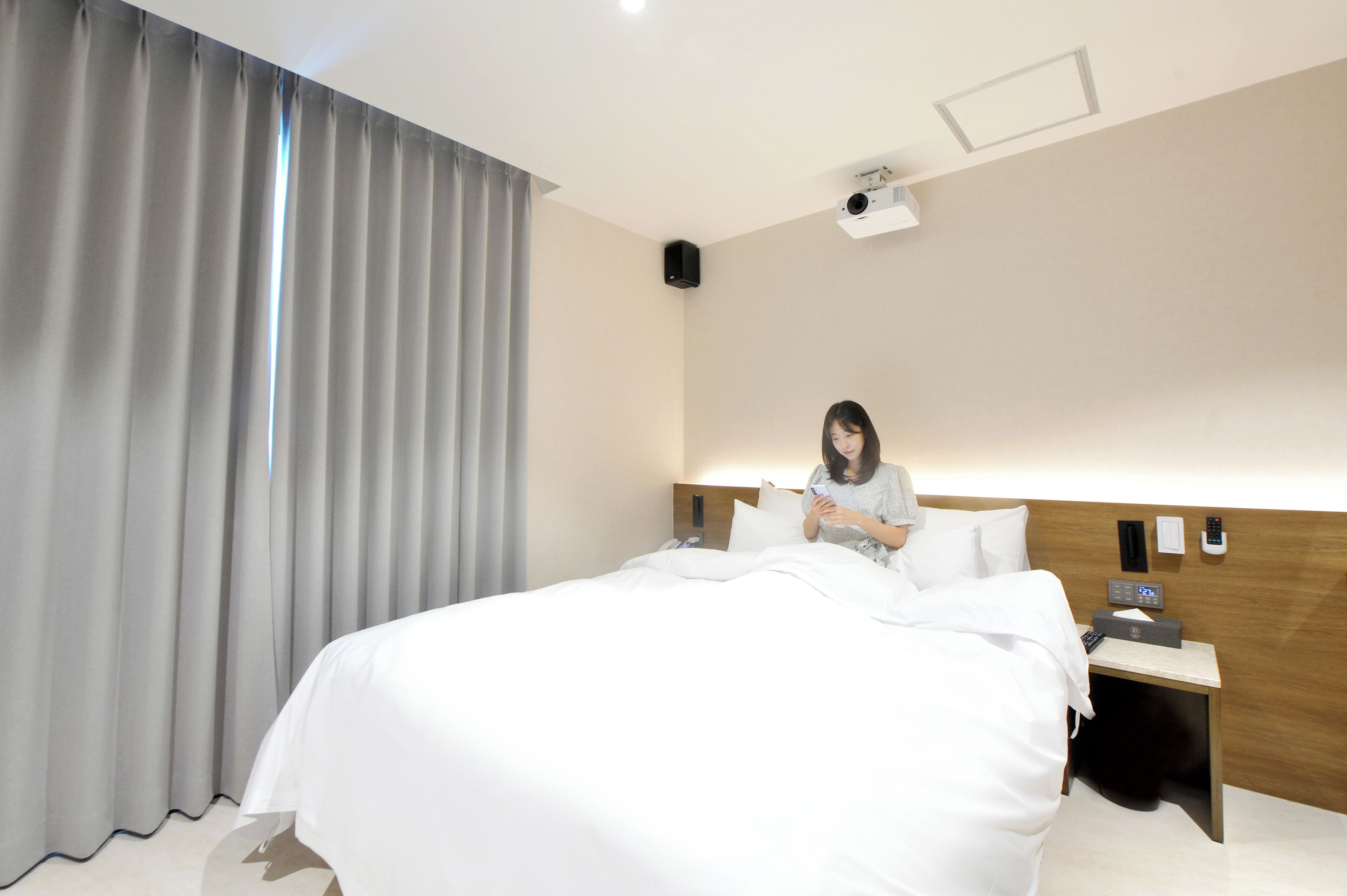 Una habitación de hotel moderna con una cama bien hecha, una mesita de noche con lámpara, un teléfono en la pared y un proyector en el techo.