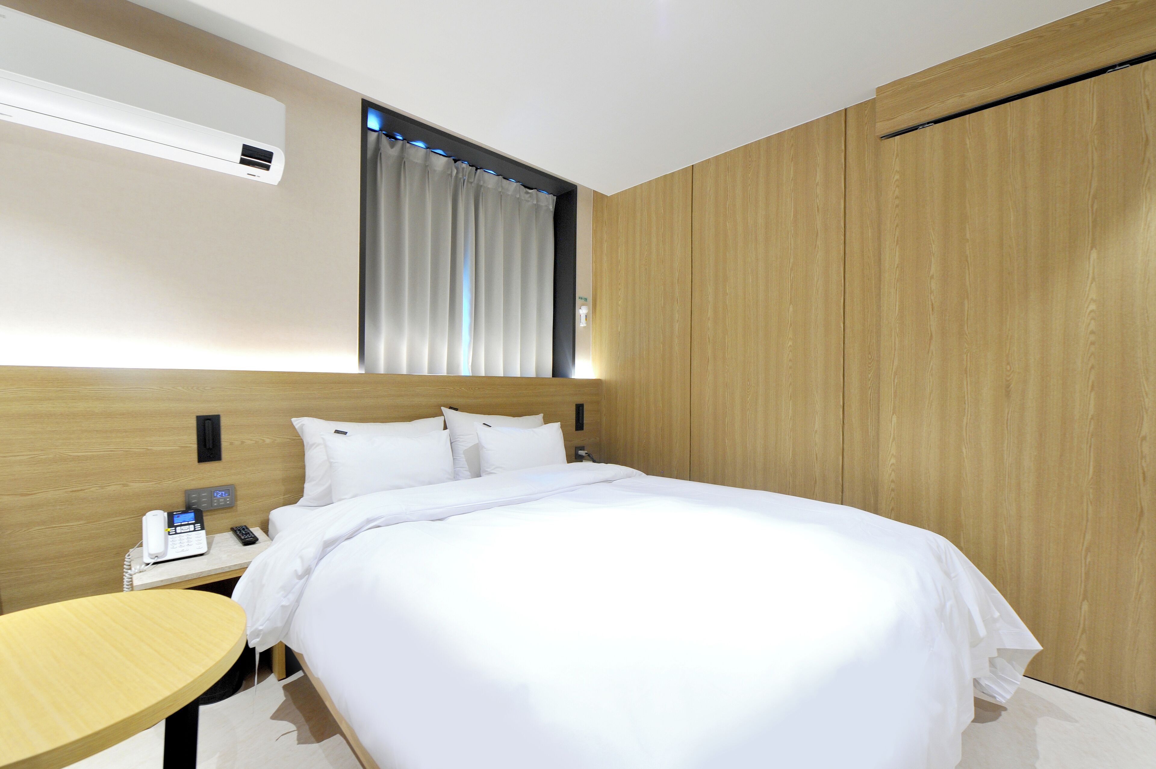 Een moderne hotelkamer met een netjes opgemaakt bed, een nachtkastje met telefoon en lamp, en een houten garderobe.