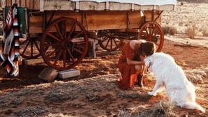 A covered wagon with wooden wheels and a white canvas roof, parked on a dirt ground with sparse vegetation.
