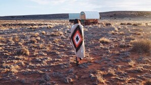 A person walking towards a covered wagon in a dry, open landscape.