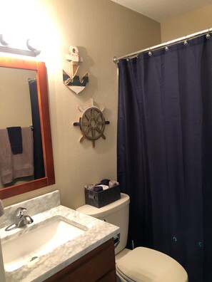 A bathroom with a white sink, a toilet, a blue shower curtain, a ship's wheel decoration, and a small storage box. - Recently Updated Waterfront Condo (Osage Beach)
