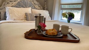 A neatly made bed with a decorative headboard, a tray with a croissant, a vase with a rose, and two mugs on a window sill.