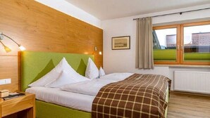Un dormitorio con cabecera verde, cama con ropa de cama blanca, manta a cuadros marrones, mesita de noche con lámpara y ventana con cortinas.