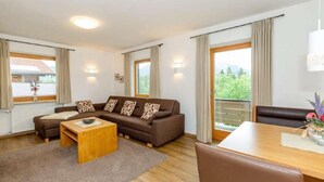 Un salón moderno con un sofá marrón, una mesa de centro de madera y una mesa de comedor con sillas. Hay ventanas grandes con cortinas, un balcón y vistas a zonas verdes.