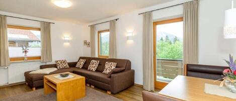 Ein modernes Wohnzimmer mit einem braunen Sofa, einem Holztisch und einem Esstisch mit Stühlen. Große Fenster mit Vorhängen, ein Balkon und Blick ins Grüne sind ebenfalls vorhanden.