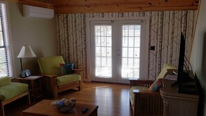 Habitació acollidora amb sostre de fusta, una finestra gran amb persianes, un sofà verd, una cadira de fusta, una taula petita amb un bol i un comandament, i una televisió sobre un suport.