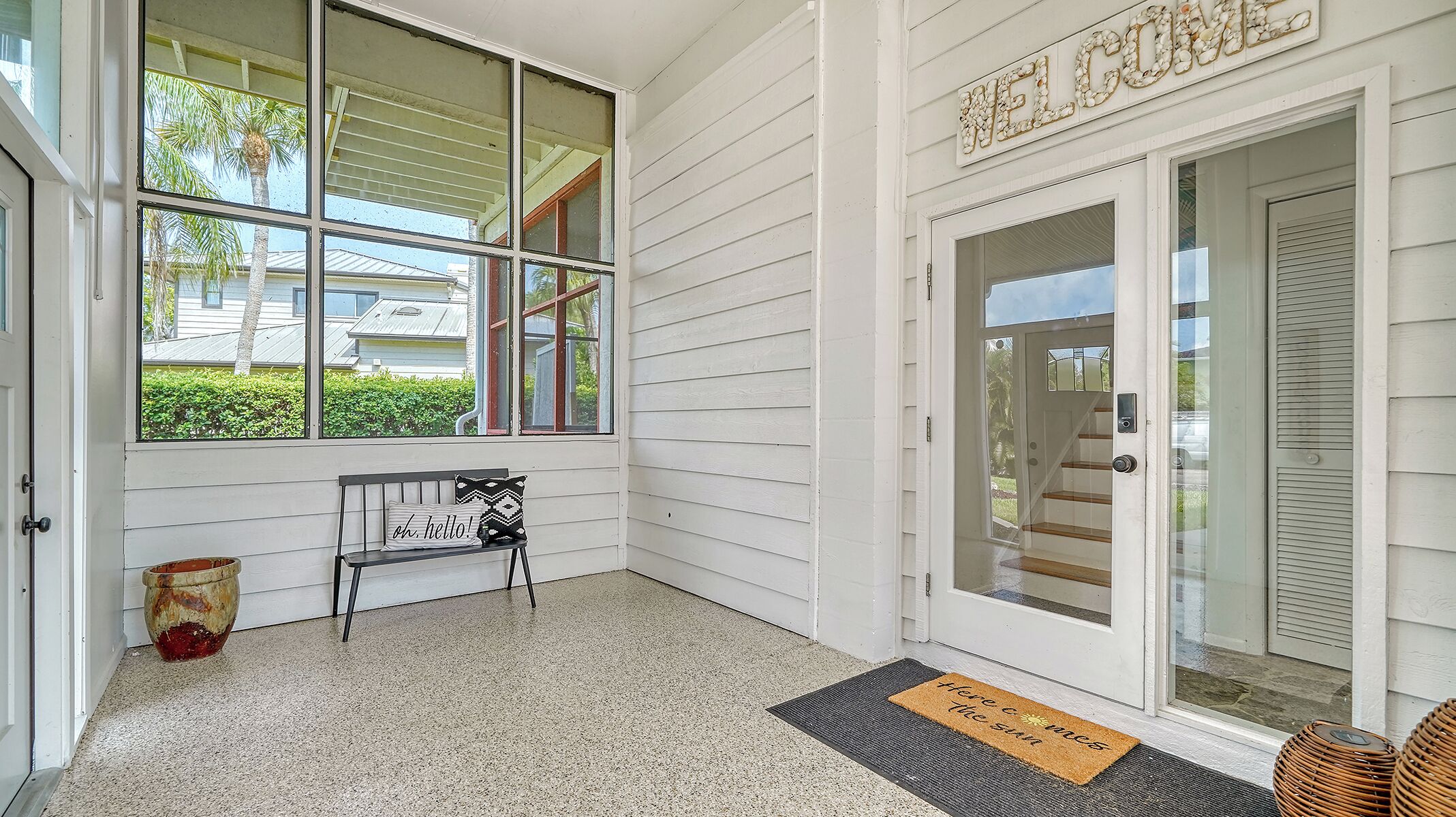 A porch with a bench and a welcome sign, a doormat, and a view of a house through the windows.