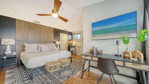 A modern bedroom with a bed, bedside tables, a desk, a chair, a ceiling fan, and a large beach painting. - Ocean View of AMI | Hot Tub | IMG | 6 TVs | 2 King Beds (Bradenton)