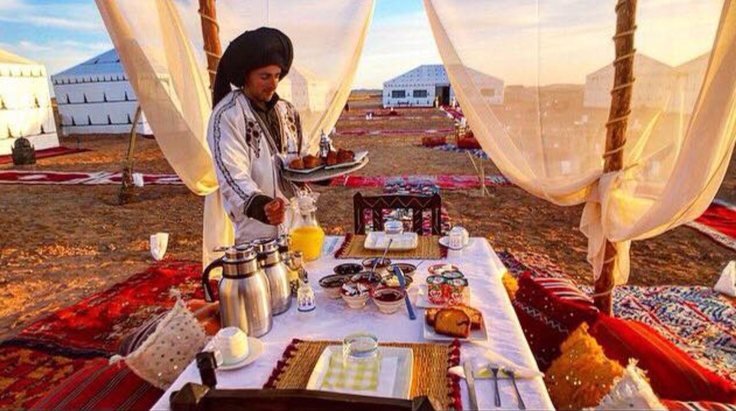 A dining setup with a table set for two, featuring a variety of dishes and drinks, under a tent with a view of yurts in the background.