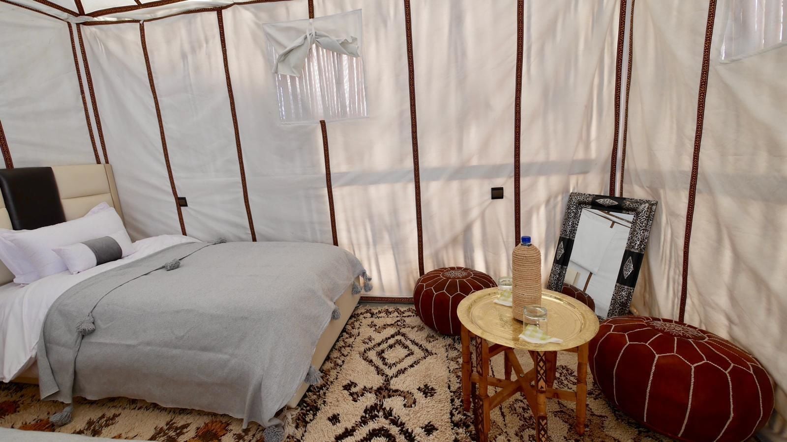 Une pièce avec un lit, une petite table ronde sur laquelle se trouve une bouteille, deux poufs rouges et un miroir au mur.