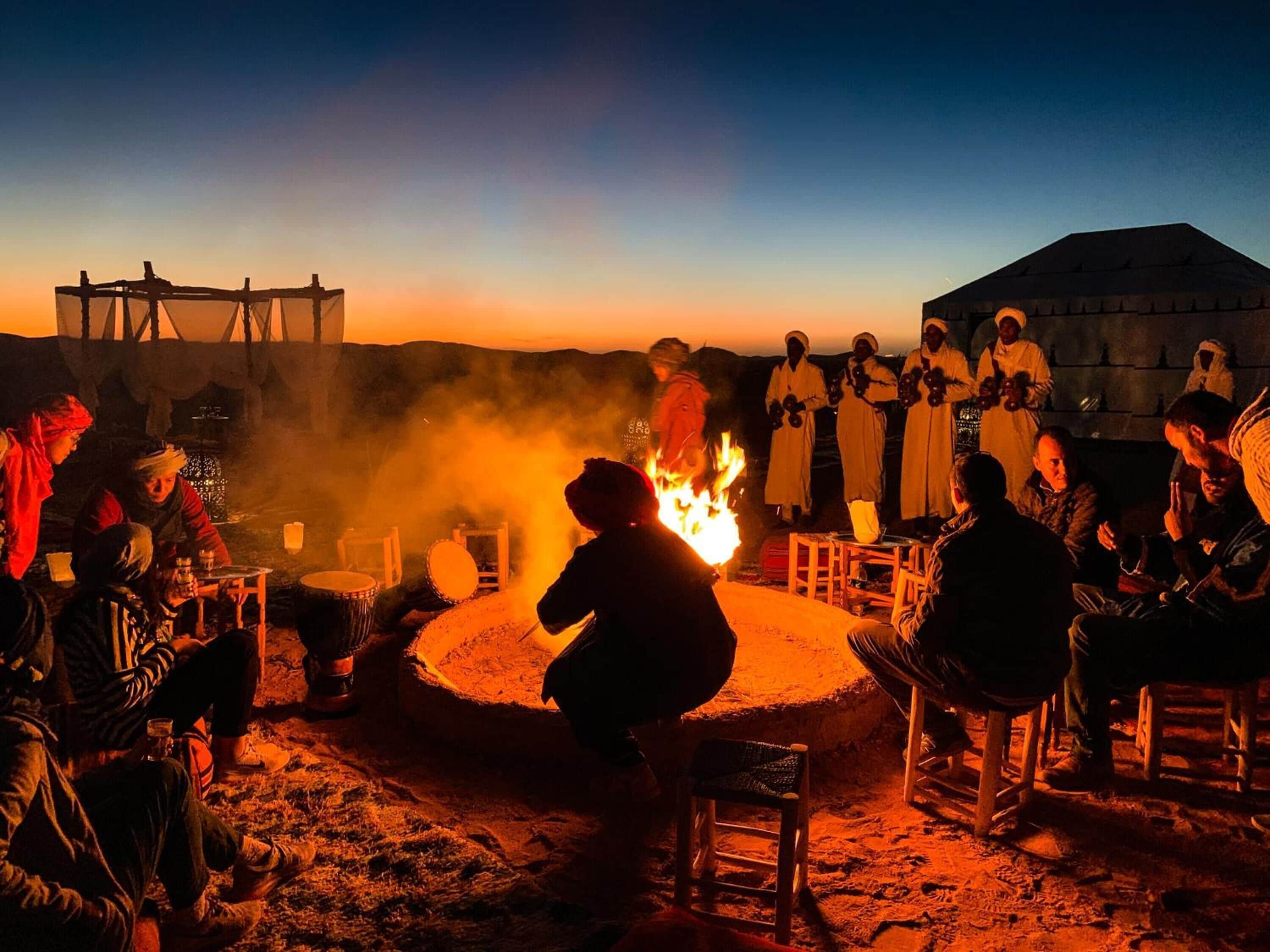 A group of people around a campfire at night, with tents and a large building in the background.
