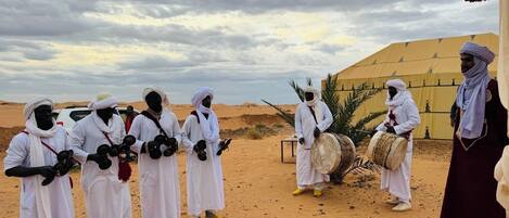 Un grupo de personas vestidas con atuendos tradicionales posando en un paisaje desértico, con una tienda de campaña al fondo.