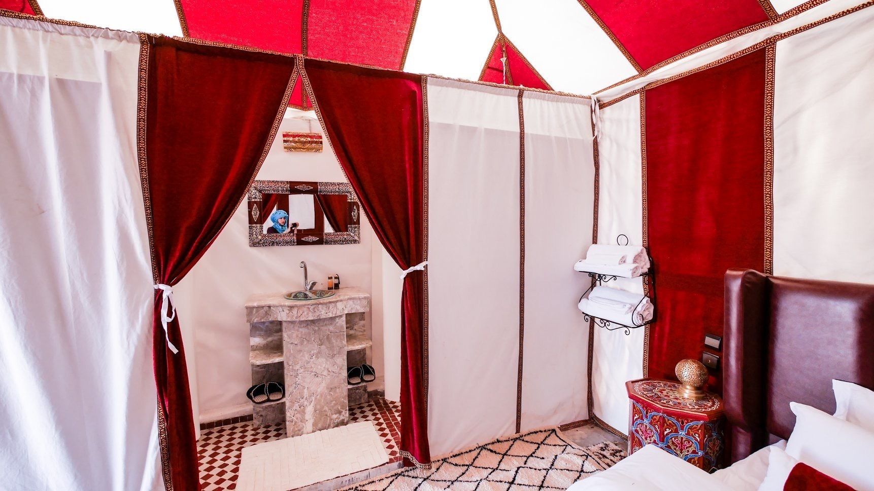 A compact hotel room with a marble sink, a small bedside table with a decorative item, and a red and white checkered floor.