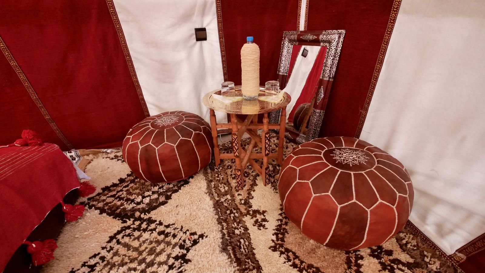 A room with two large, round ottomans with a geometric pattern, a small wooden table with a bottle and glass, and a mirror on the wall.